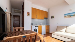 A compact living space with a dining table, wooden chairs, a sofa, a kitchen area with wooden cabinets, a washing machine, and a small sink.