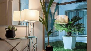 Un intérieur moderne avec une grande fenêtre, une table en verre surmontée d’une lampe et deux plantes en pot.