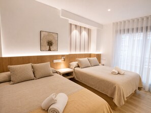 A hotel room with two beds, a nightstand, a wall-mounted light, and a window with sheer curtains.