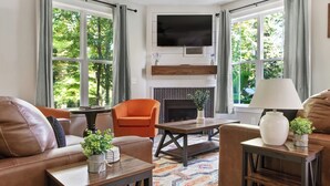 Una sala de estar con chimenea, un televisor montado encima y un sofá. Hay dos ventanas con cortinas, y una mesa con una lámpara y una planta.