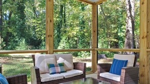 Una habitación luminosa con muebles de mimbre, ventanas grandes y vistas a un bosque.