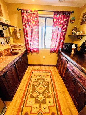 A narrow kitchen with wooden cabinets, a patterned rug, and a window with curtains. - Romeo's Roadhouse with Cowboy Pool (Lake Montezuma)