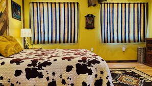 A room with a cowhide-patterned bedspread, two windows with striped curtains, a wall-mounted lamp, and a rustic decor including a hat and a wooden guitar. - Romeo's Roadhouse with Cowboy Pool (Lake Montezuma)