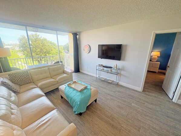 Un salon avec un canapé beige, une table basse en verre, un téléviseur fixé au mur et un balcon donnant sur un paysage verdoyant.