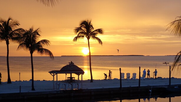 A beach scene with a thatched hut, palm trees, and calm waters reflecting the sunset. - Charming Bayside 2 bed, 2 bath Condo at Futura in Islamorada WiFi, Pool, Dockage (Tavernier)