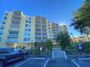 Un immeuble résidentiel à plusieurs étages, doté de balcons, d’un espace de stationnement et d’un chemin menant à l’entrée.
