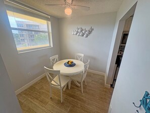 Un coin repas avec une table ronde, des chaises blanches, un ventilateur de plafond et une fenêtre donnant sur des immeubles.
