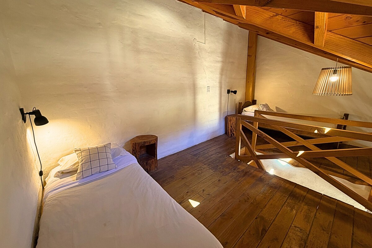 A room with a wooden bed, a small stool, a sloped ceiling, and a wall-mounted lamp.