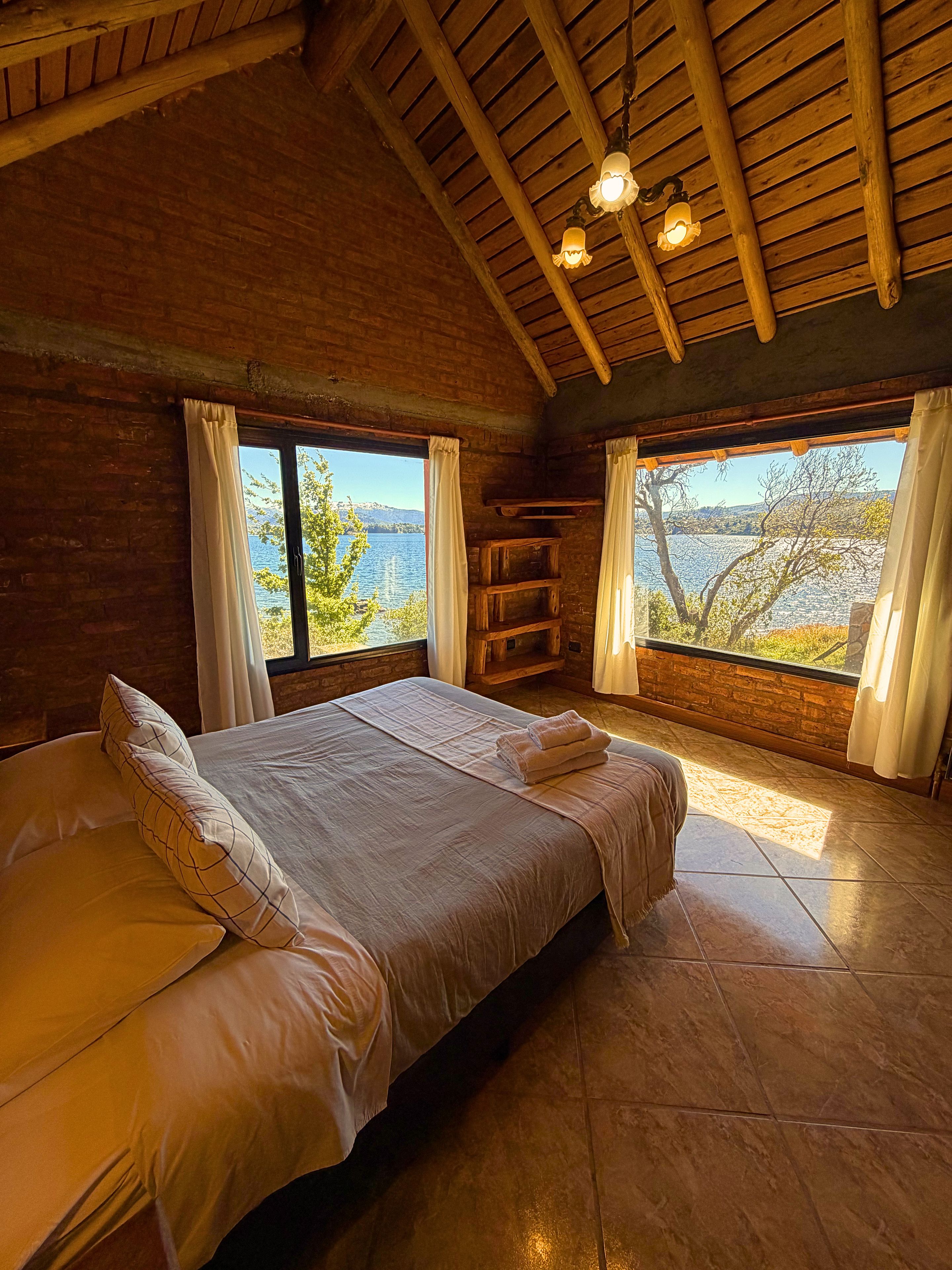 A bedroom with a large bed, two windows with curtains, a wooden ceiling, and a view of the outdoors.