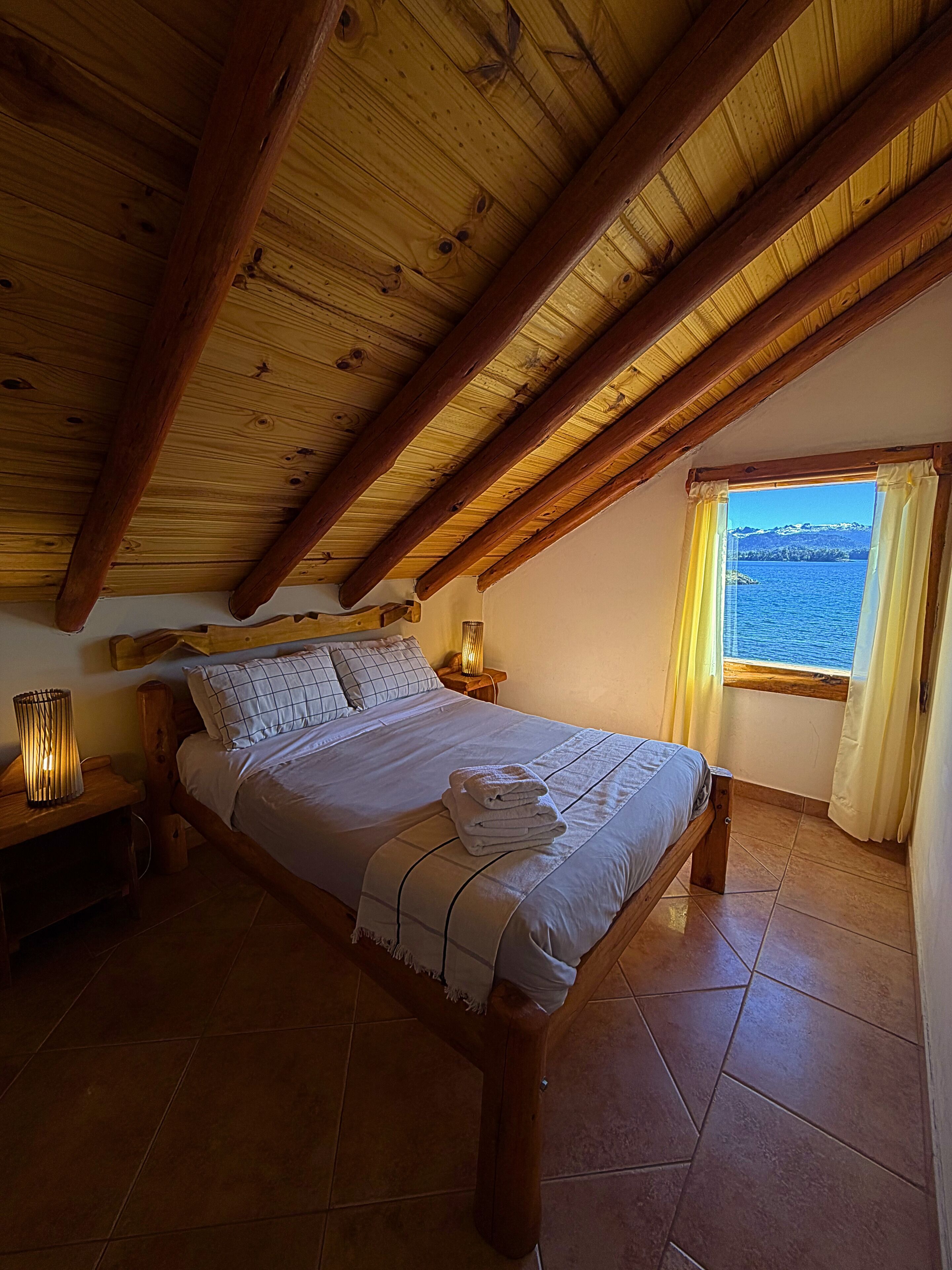 A bedroom with a wooden ceiling, a bed with a white bedspread, a nightstand with a lamp, and a window with a view of the sea.
