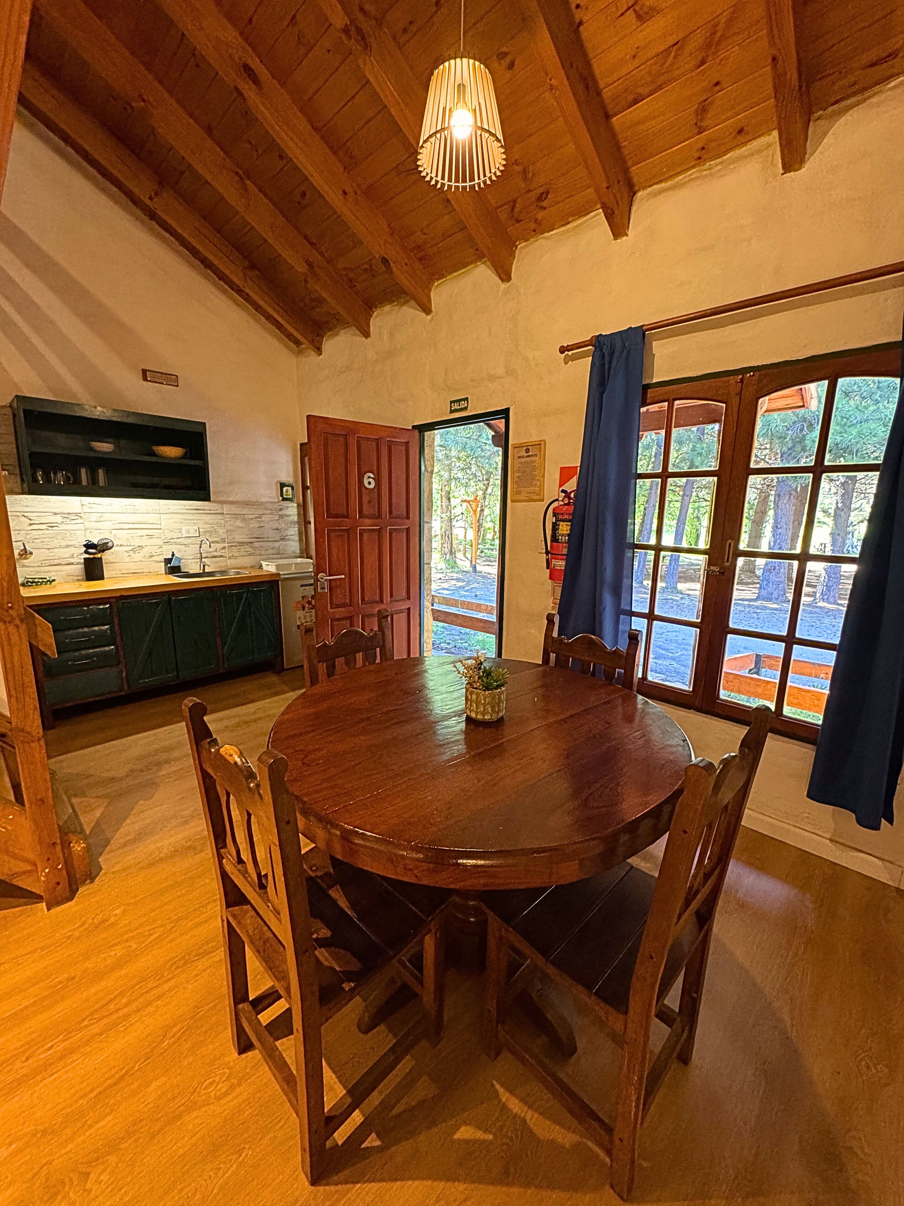 Taula de dinar de fusta amb cadires, zona de cuina amb un rentaplat i armaris, i una porta que dona a l'exterior.