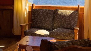 Une cabane avec un intérieur en bois, un canapé agrémenté de coussins à motifs, une table en bois et une fenêtre avec des rideaux donnant sur un lac et des montagnes.