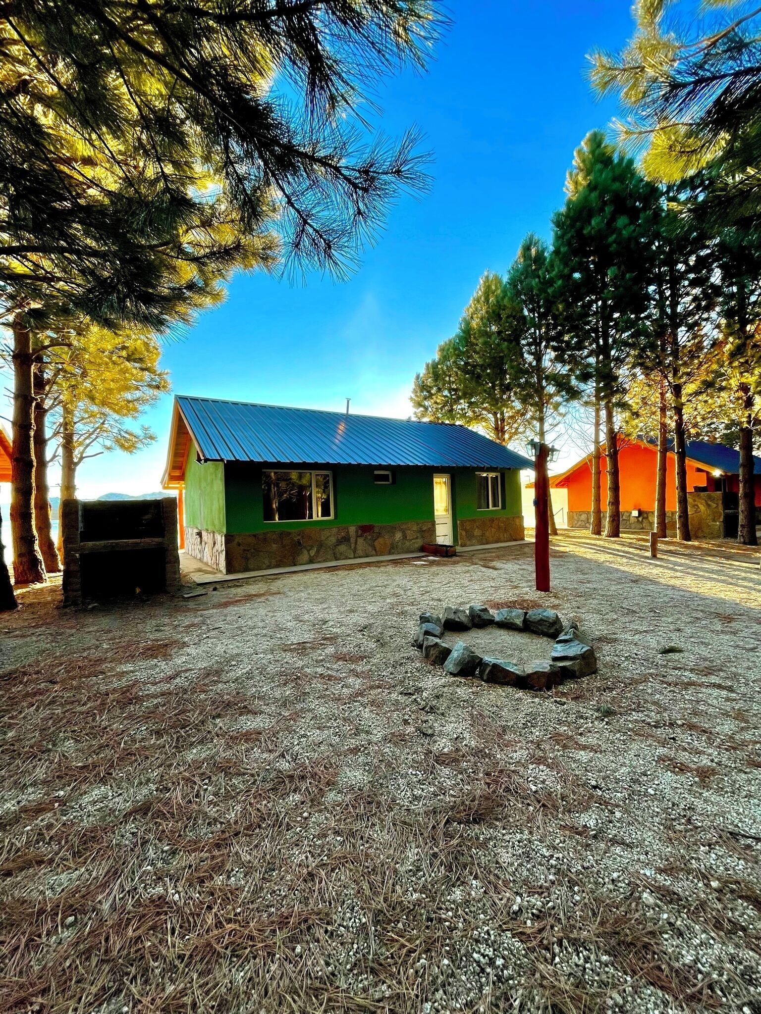 Un edificio de fachada verde y techo azul, rodeado de árboles y un área de grava con un hogar de piedra.