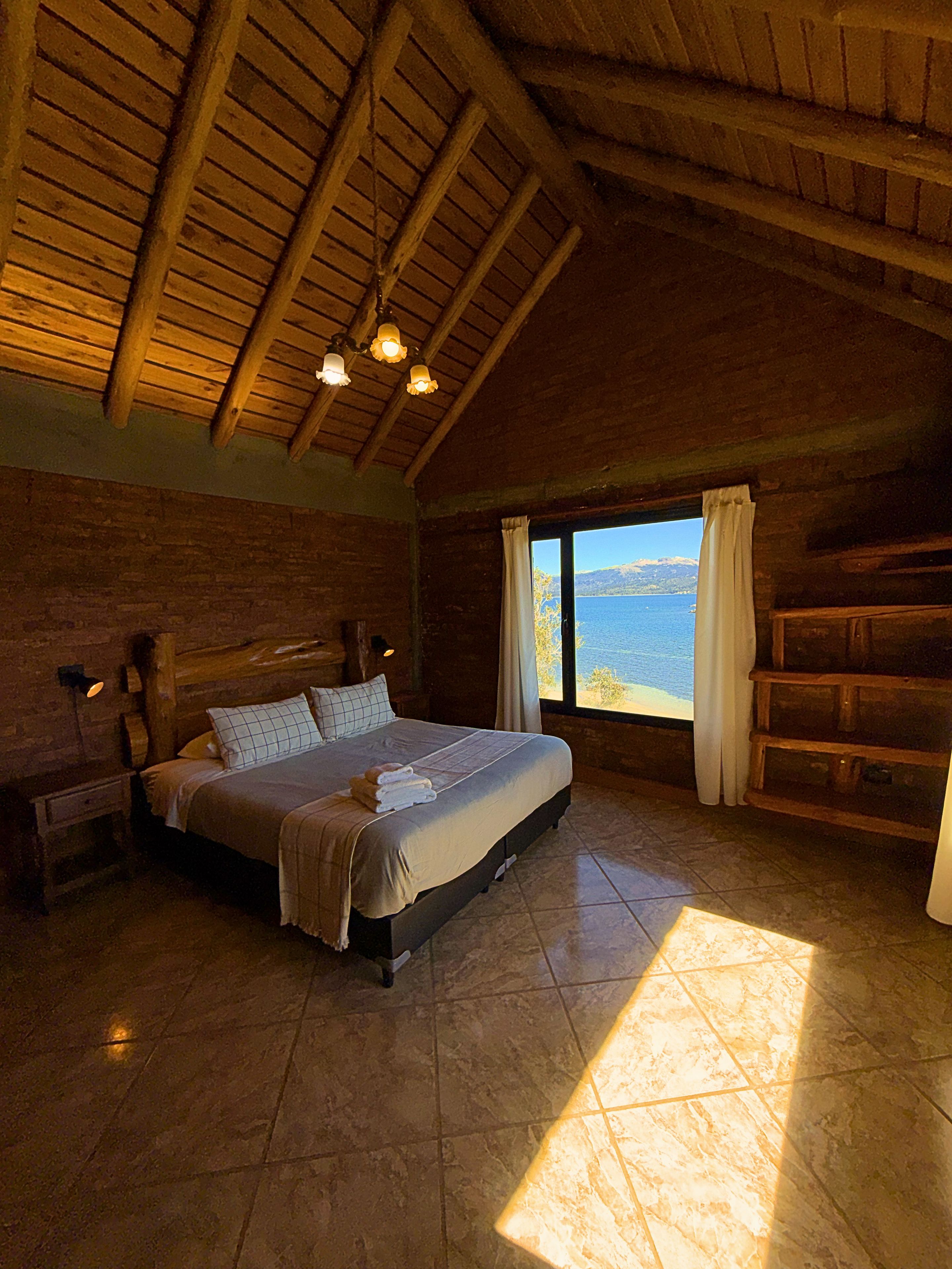 Une chambre avec un grand lit, des poutres apparentes, une table de chevet, une fenêtre avec vue et un banc intégré.