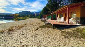Une propriété située en bord de mer, dotée d’une terrasse en bois, de chaises longues et offrant une vue sur l’eau et les montagnes.