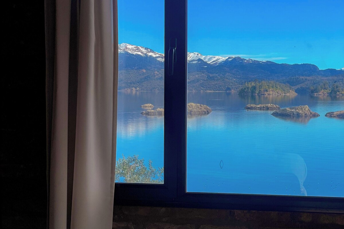 A hotel room with a bed, a window with a view of a lake and mountains, and a stack of towels on the bed.