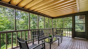A covered outdoor deck with wooden flooring and metal railings, featuring two black chairs and a small table with a plant. - Updated, Cozy home- minutes to Tunnel Vision Trail!  (Bella Vista)