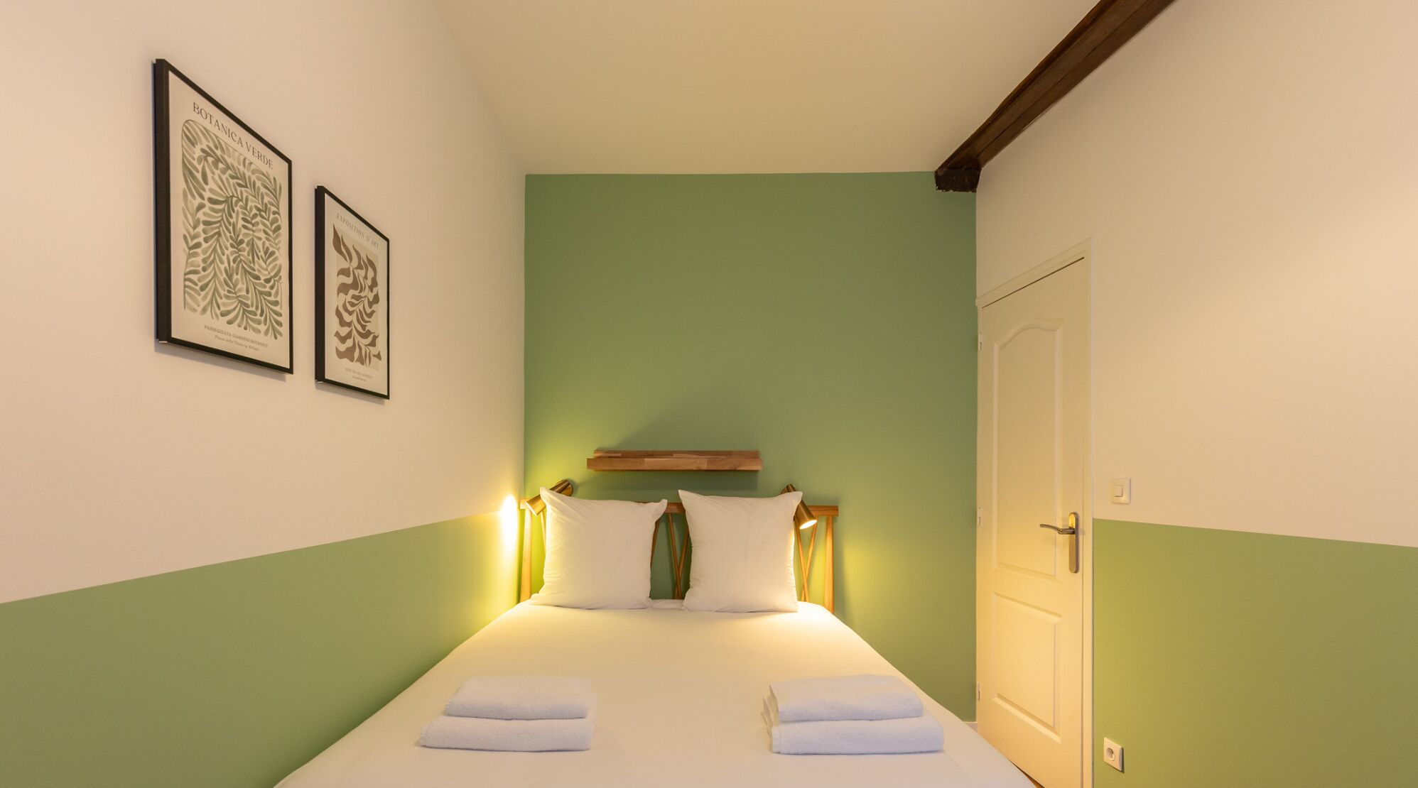 Une petite chambre à coucher, bien rangée, avec un lit, deux reproductions botaniques encadrées et une porte.
