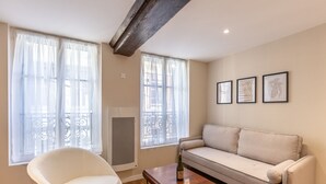 A living room with a sofa, a chair, and a table. There are two windows with curtains, a balcony railing, and three framed botanical prints on the wall.