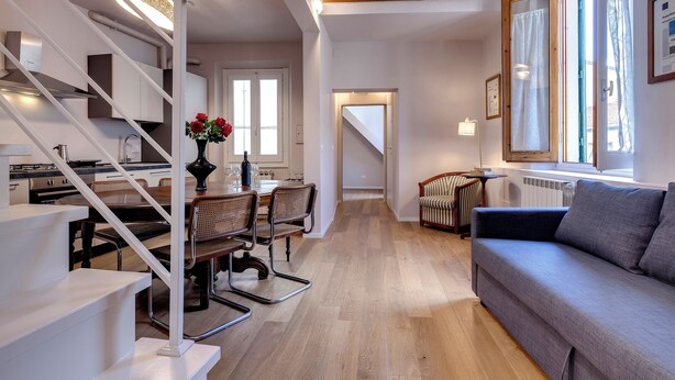 Um interior moderno com uma escada que leva a um quarto em mezanino, uma sala de jantar com mesa e cadeiras, e uma sala de estar com sofá e janela.