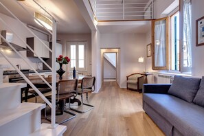 Et moderne interiør med en trappe op til et loftssoveværelse, et spiseområde med bord og stole, og en opholdsstue med sofa og et vindue.