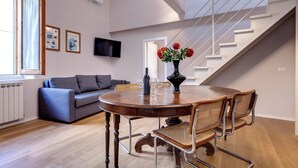 Uma sala de estar moderna com uma mesa de jantar de madeira, cadeiras de vime, um sofá cinza, uma escada e uma TV fixada na parede.