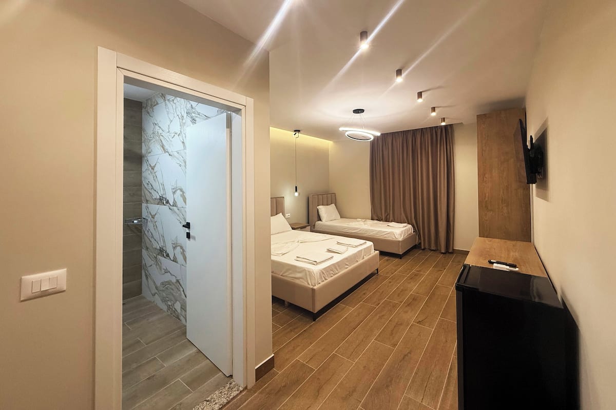 Una habitación de hotel moderna con piso de madera, dos camas, un televisor y un baño con baldosas de mármol.