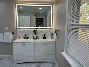 Un baño con tocador blanco, un espejo grande y una ventana con persianas.
