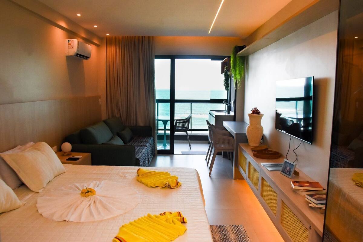 A hotel room with a bed, a desk, a TV, and a balcony with a view of the sea.