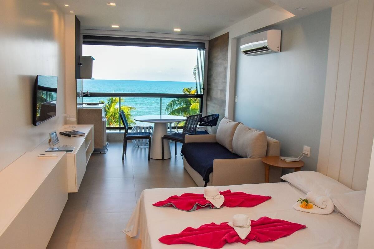 A hotel room with a bed, a desk, a chair, a sofa, and a balcony with a view of the sea.