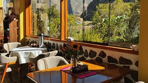 A dining area with a table, chairs, and a view of mountains through large windows. - Sara Garden Lodge (Ollantaytambo)