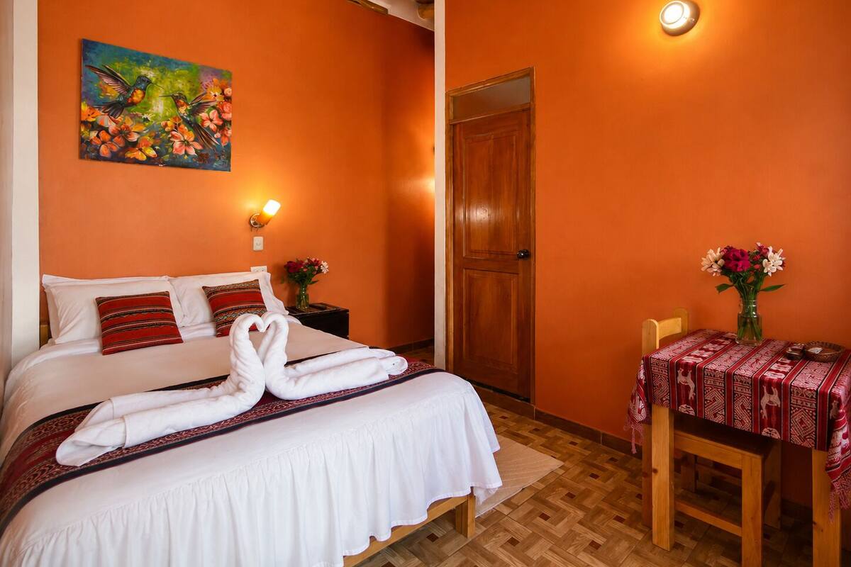 A hotel room with a bed, a painting, a door, a table with a tablecloth, and a vase with flowers.