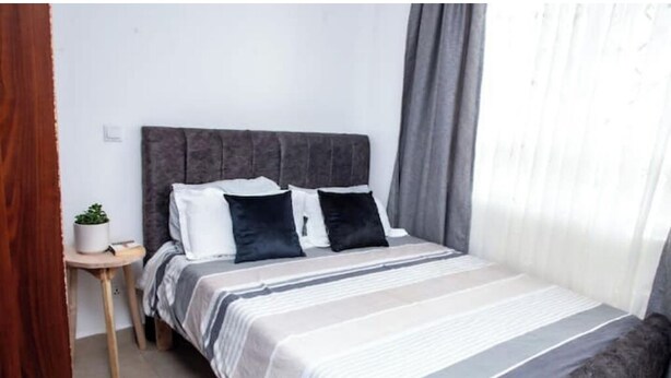 A neatly made bed with a striped bedspread and two pillows, a small wooden side table with a plant, and a window with curtains.