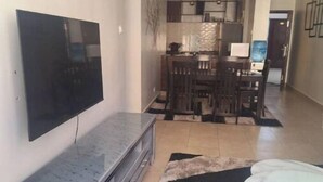 A modern living room with a flat-screen TV mounted on the wall, a grey entertainment unit, a dining area with a dark wooden table and chairs, and a patterned rug on the floor.
