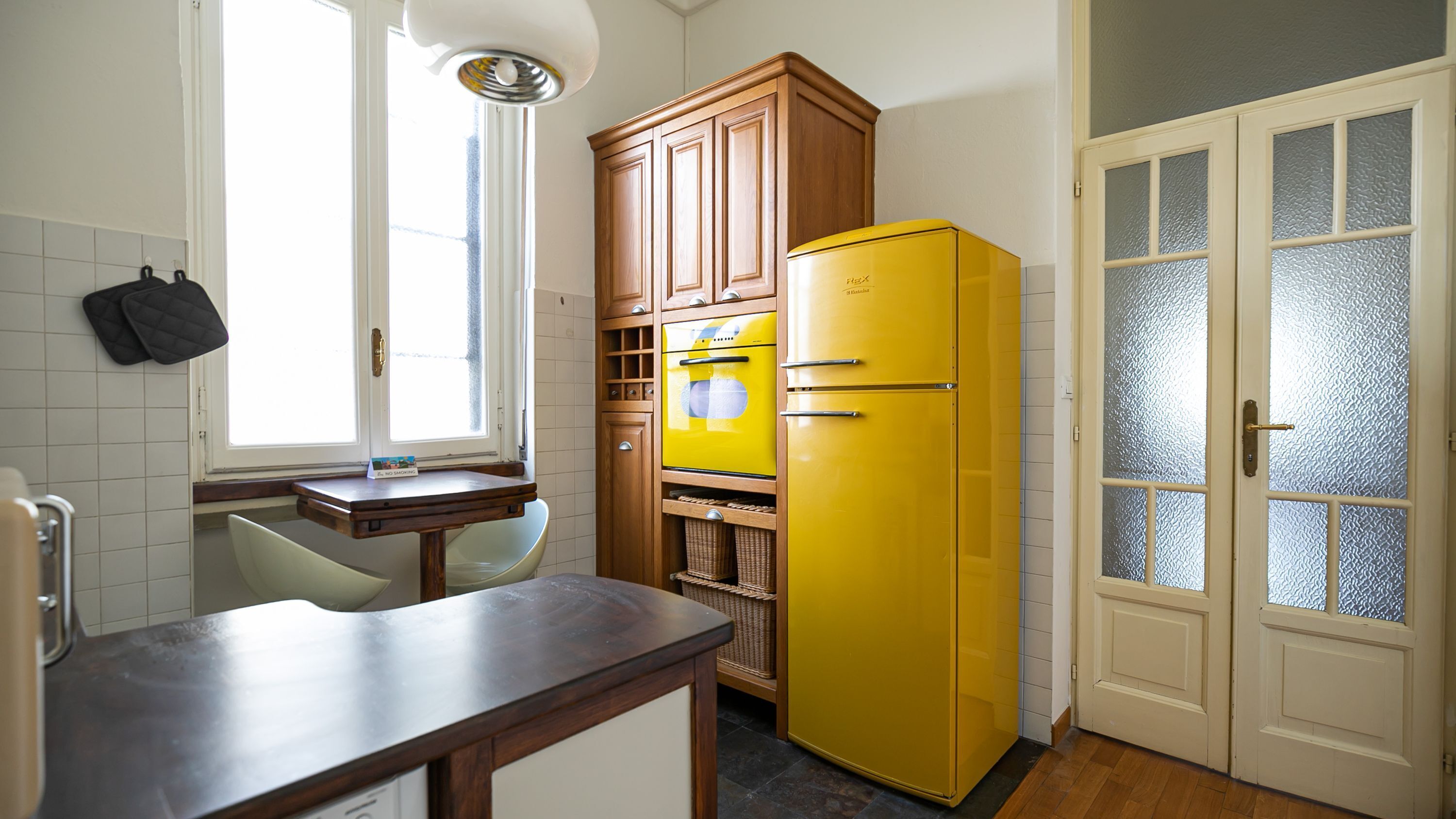 A kitchen with a wooden cabinet, a yellow refrigerator, a window with blinds, and a door with frosted glass panels.