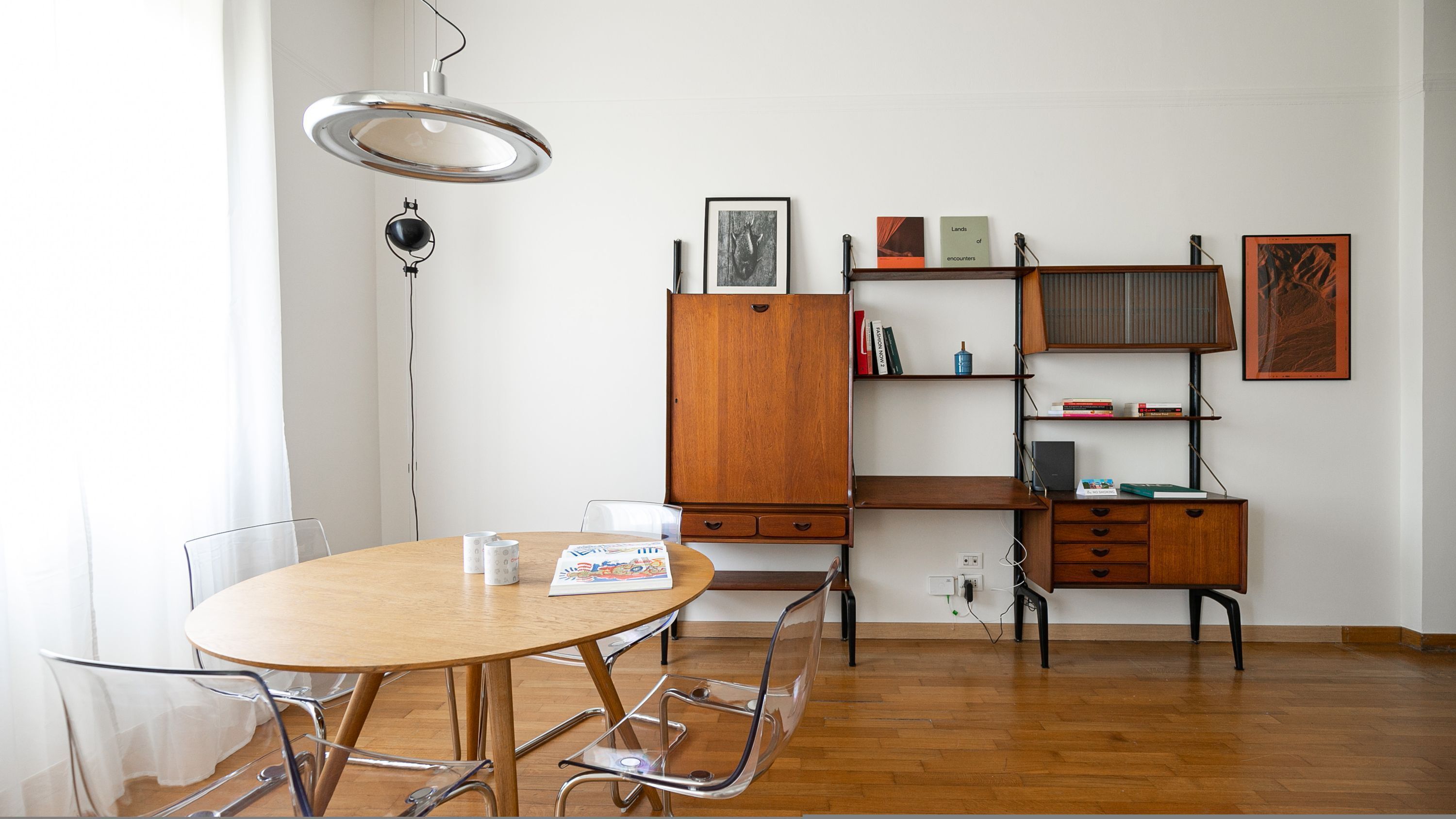 A room with a wooden dining table, chairs, a cabinet, and shelves with books and framed pictures.