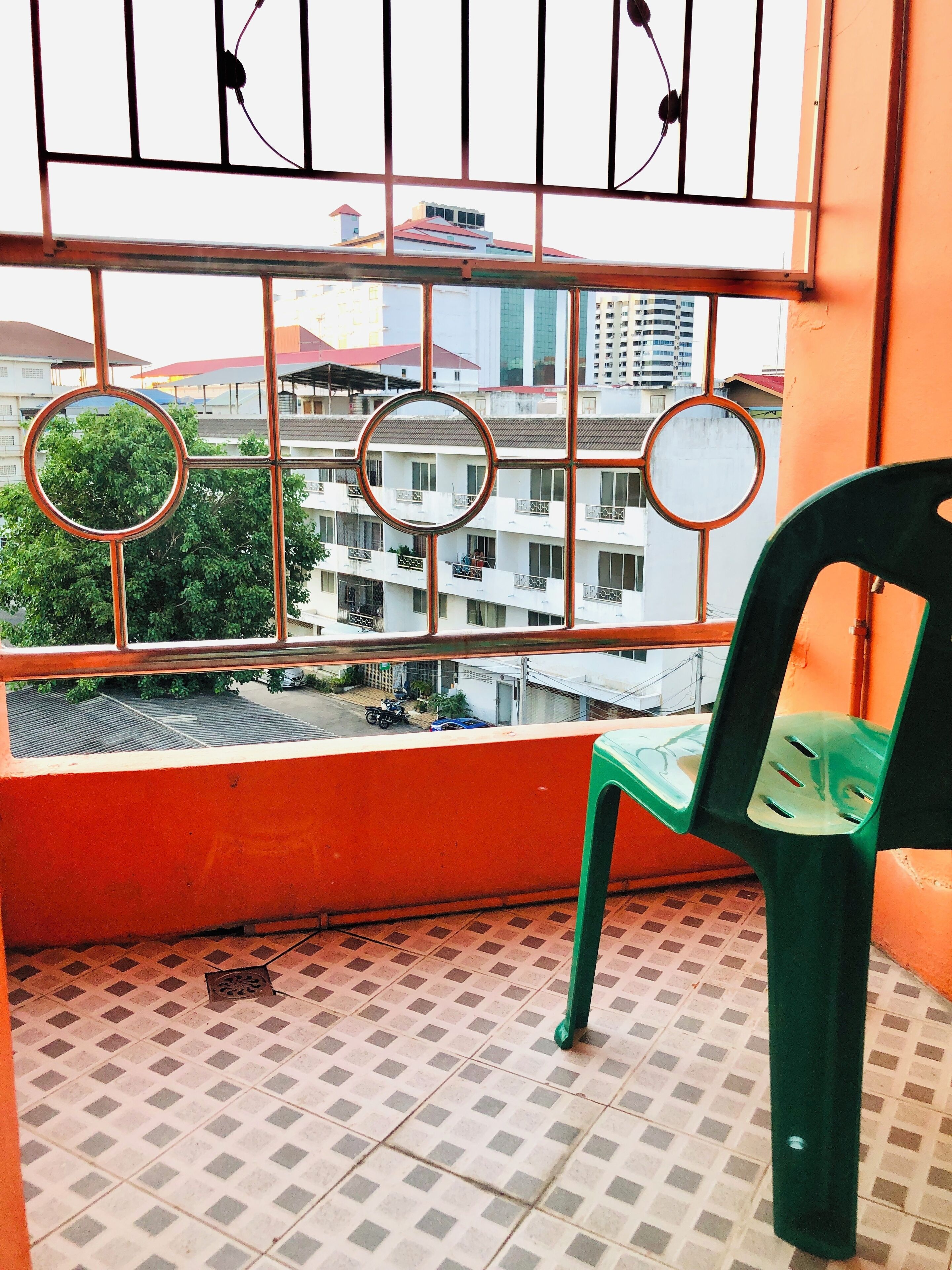 A balcony with a view of buildings, a chair, and a window with circular cutouts.