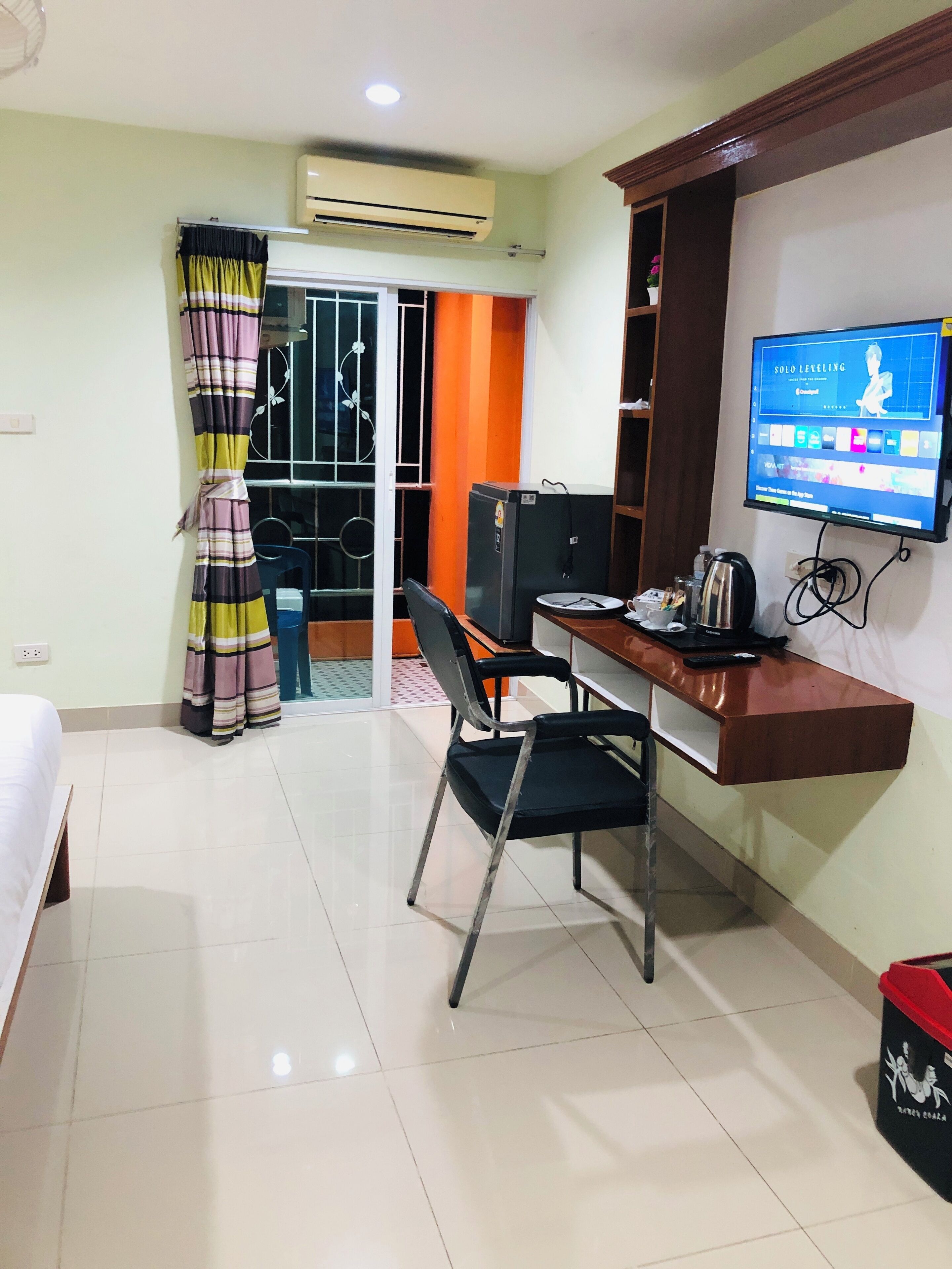 A hotel room with a bed, a desk with a chair, a television, an air conditioner, and a red trash bin.