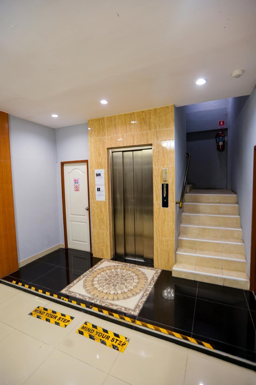 A hotel lobby with a central elevator, a staircase, and a patterned floor mat.