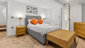 A modern bedroom with a grey bed, orange pillows, a wicker bench, and a wooden dresser.
