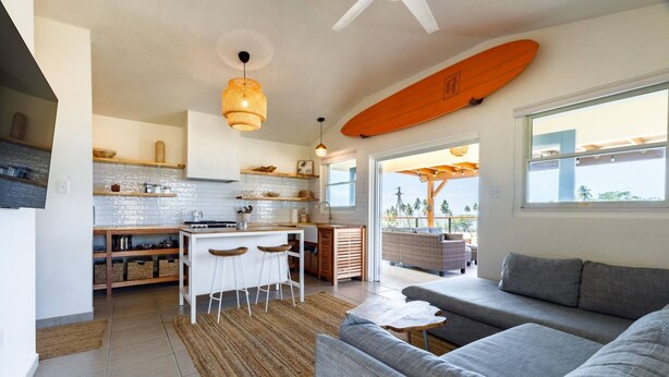 Une cuisine moderne avec des meubles blancs, un îlot central doté de tabourets et un coin repas avec un canapé en osier et une vue sur l’extérieur.