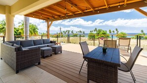 Un espace extérieur aménagé, abrité et doté d’une terrasse en bois, d’un canapé, d’une table et de chaises, offrant une vue sur une plage bordée de palmiers et sur l’océan.