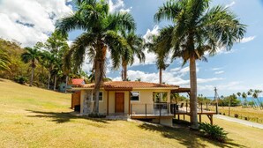Une petite maison de plain-pied, avec un toit en tuiles, entourée de palmiers et d’une pelouse.
