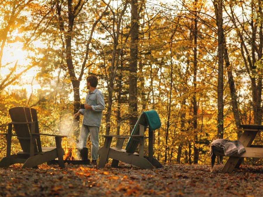 En person sitter vid en lägereld i en skog, med två stolar och ett picknickbord i närheten.