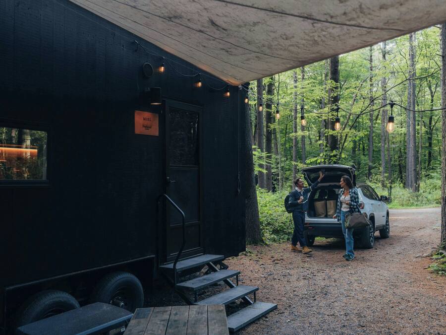 A black RV with a covered porch, steps leading to a door, and a car parked nearby.