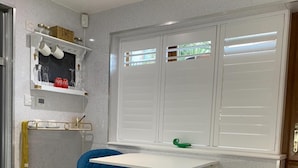 Une cuisine compacte avec un tabouret de bar bleu, un plan de travail blanc et une fenêtre avec des stores.