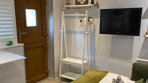 Une chambre à coucher avec un lit, un téléviseur fixé au mur, une porte en bois, une étagère blanche et une petite fenêtre.