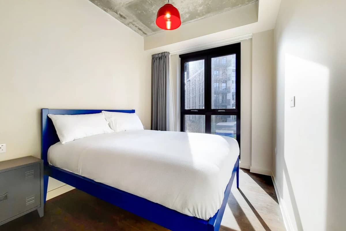 Quarto moderno com estrutura de cama azul, roupa de cama branca, luminária pendente vermelha e janela com cortinas.
