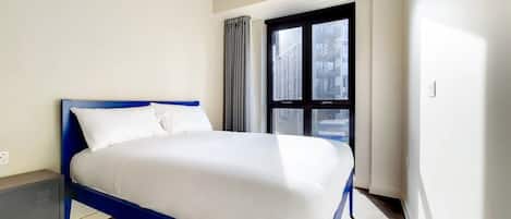 A modern bedroom with a blue bed frame, white bedding, a red pendant light, and a window with curtains.