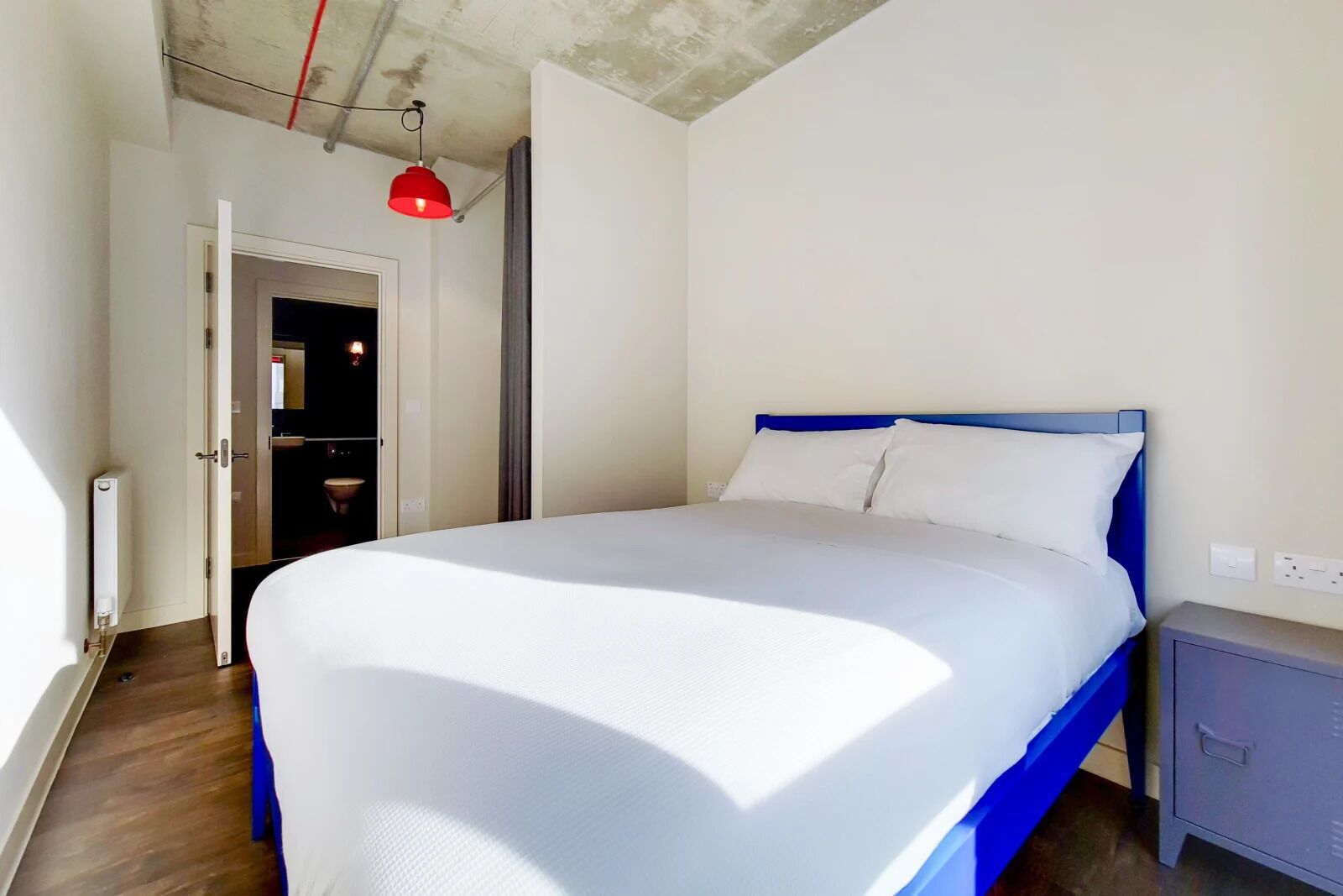Quarto com cama, mesa de cabeceira, luminária pendente vermelha e porta de acesso a um banheiro.
